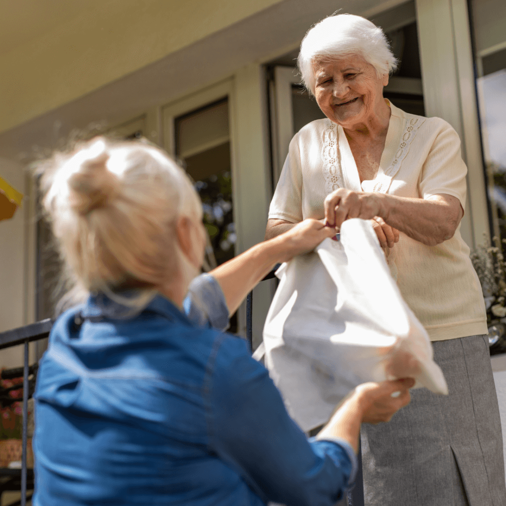 A blonde woman hands a bag to an elderly woman with short white hair, standing on a balcony. - Home Instead