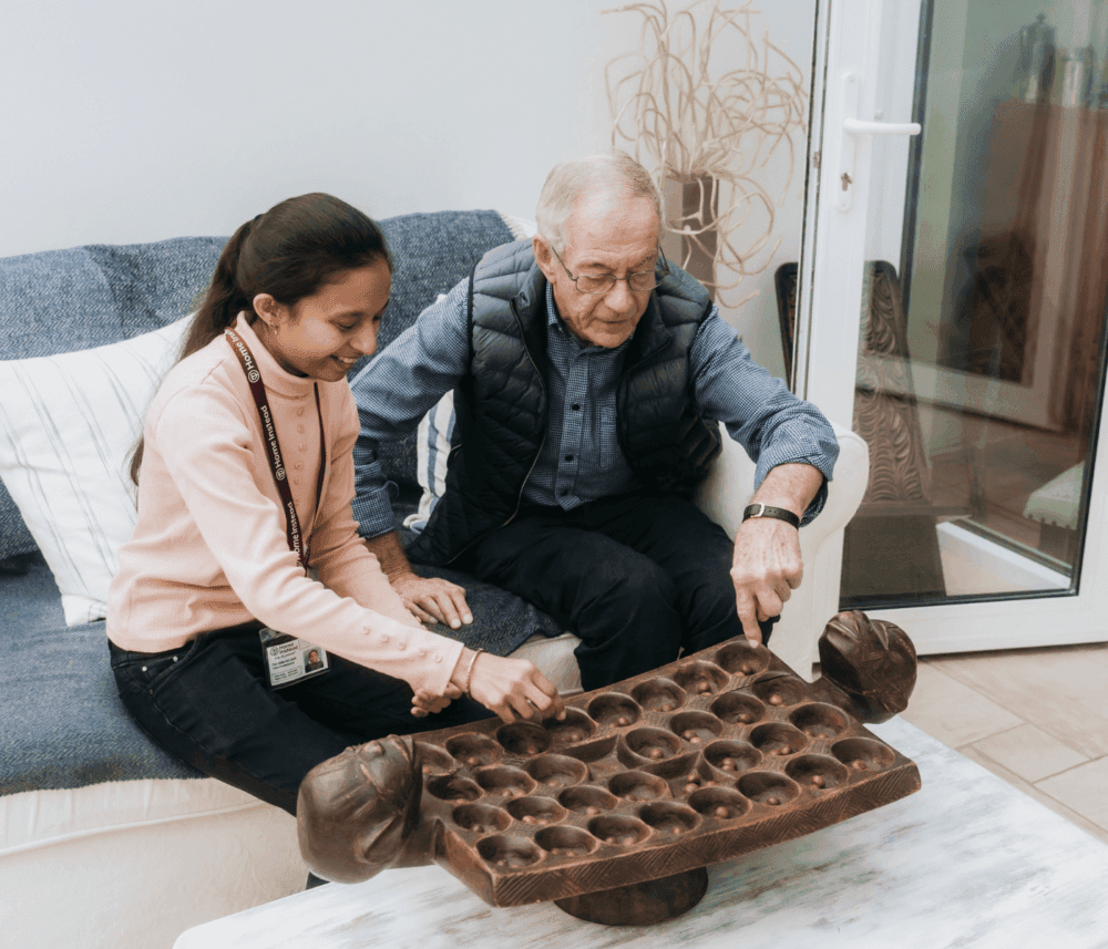 An elderly man and a young girl playing a game on a carved wooden board while sitting on a couch. - Home Instead