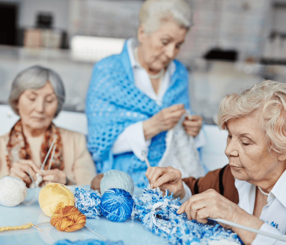 Three elderly women knitting and crocheting with colorful yarns, sitting at a table intently focused on their work. - Home Instead