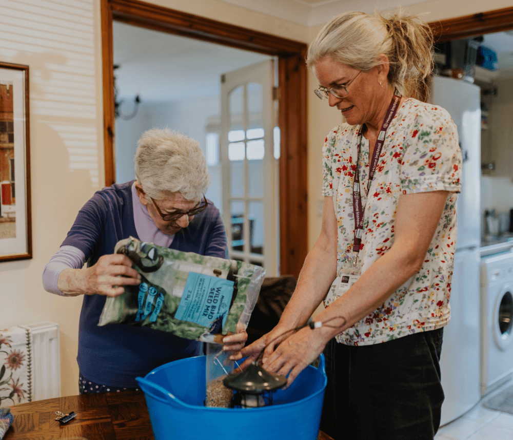 Two women, one elderly and one middle-aged, preparing bird food in a home by pouring seeds into a large blue container. - Home Instead