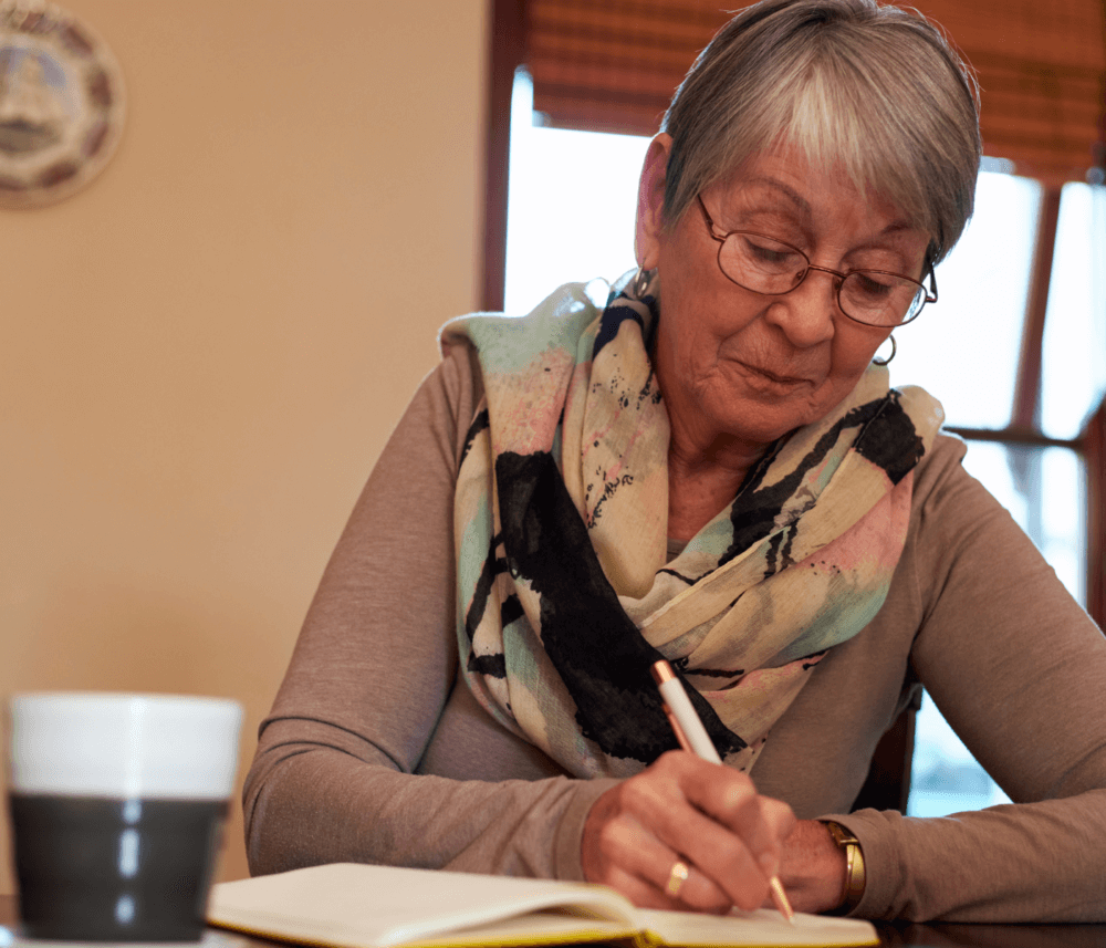 An elderly woman with glasses and a scarf writes in a notebook at a table beside a coffee cup. - Home Instead