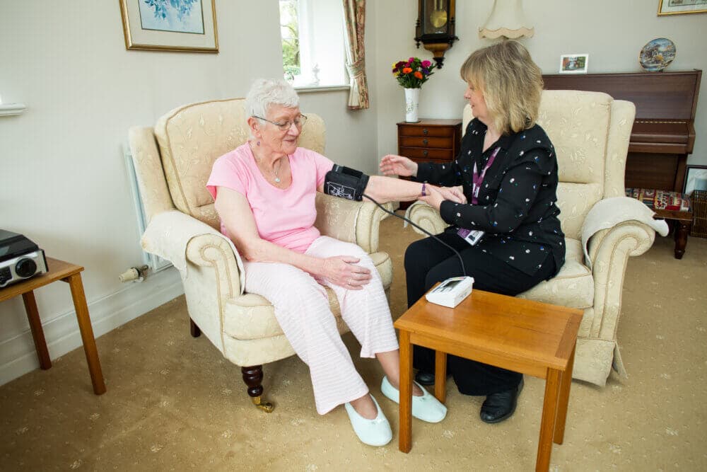 A woman checks the blood pressure of an elderly woman sitting in an armchair in a cozy living room. - Home Instead