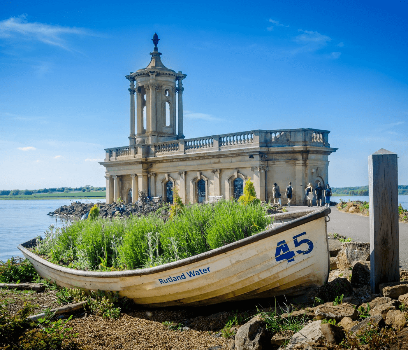 Old boat with plants and "Rutland Water 45" written on it, in front of a lakeside church under a clear blue sky. - Home Instead