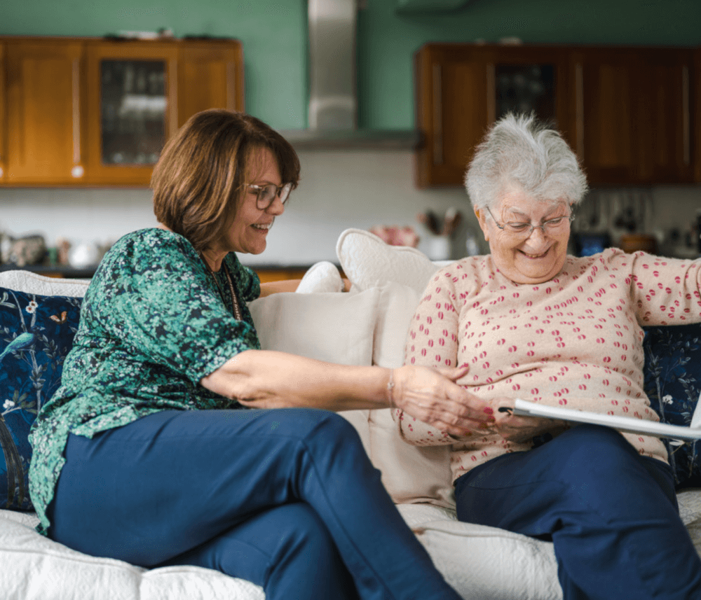 Two women sitting on a couch, smiling, and looking at a photo album together in a cozy living room. - Home Instead