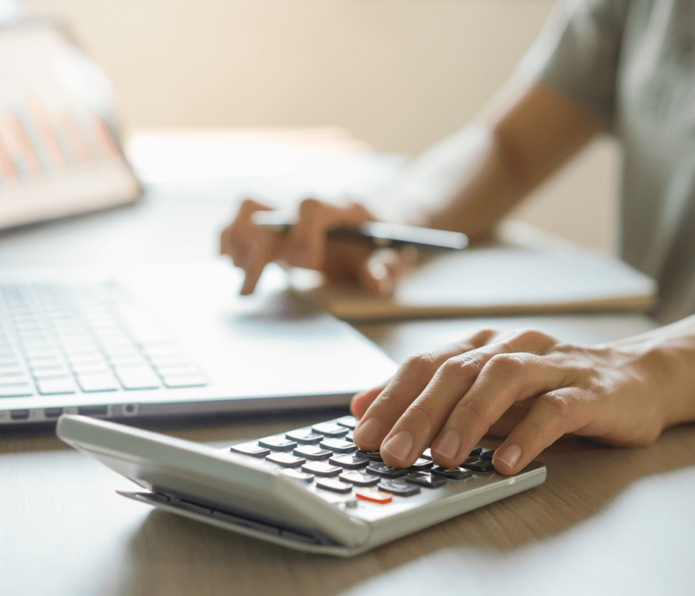Person using a calculator and taking notes while working on a laptop at a desk. - Home Instead