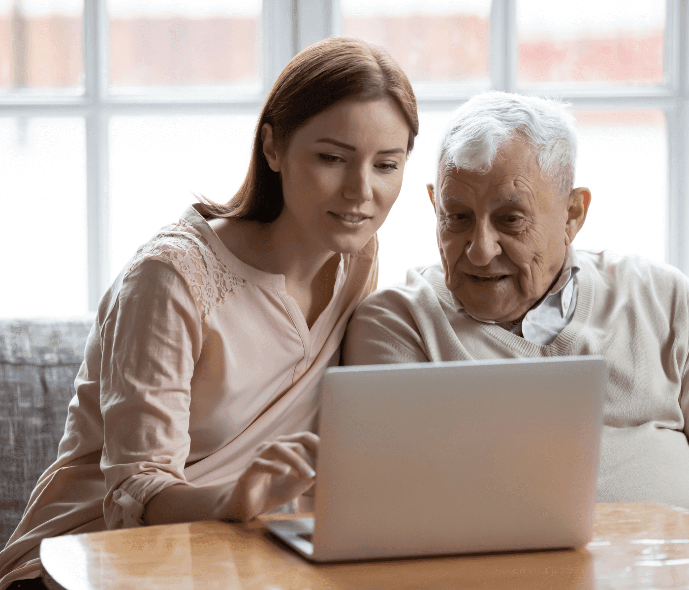 A young woman helps an elderly man use a laptop while sitting together at a table in a bright room. - Home Instead