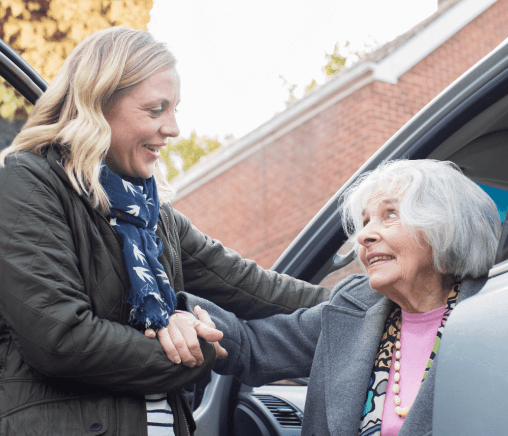 A woman helps an elderly woman out of a car, smiling and holding hands, outside on a sunny day. - Home Instead