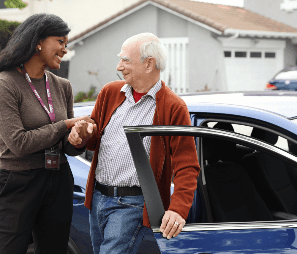 A woman helps an elderly man out of a car in a residential area, both smiling. - Home Instead