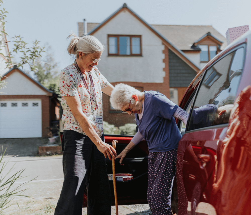A woman helps an elderly person with a walking cane out of a red car in a suburban neighborhood. - Home Instead