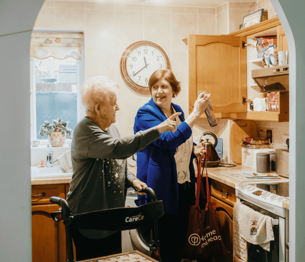 An elderly woman and a caregiver happily organize a kitchen together, with the caregiver holding a shopping bag. - Home Instead