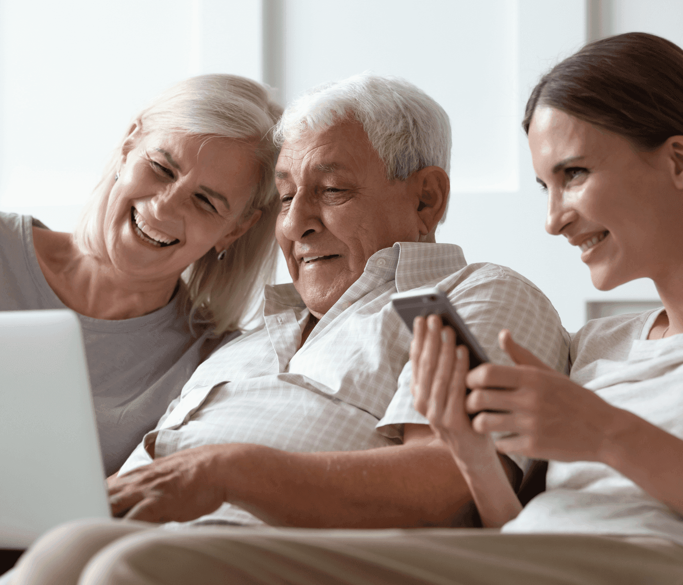 Three people smiling, sitting on a couch. An older man looks at a laptop with two women, one of whom is holding a phone. - Home Instead