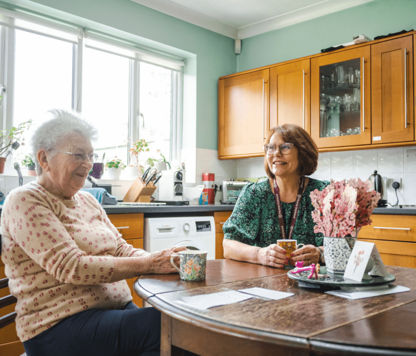 Two women sitting at a kitchen table, smiling and drinking tea, with flowers and cards on the table. - Home Instead