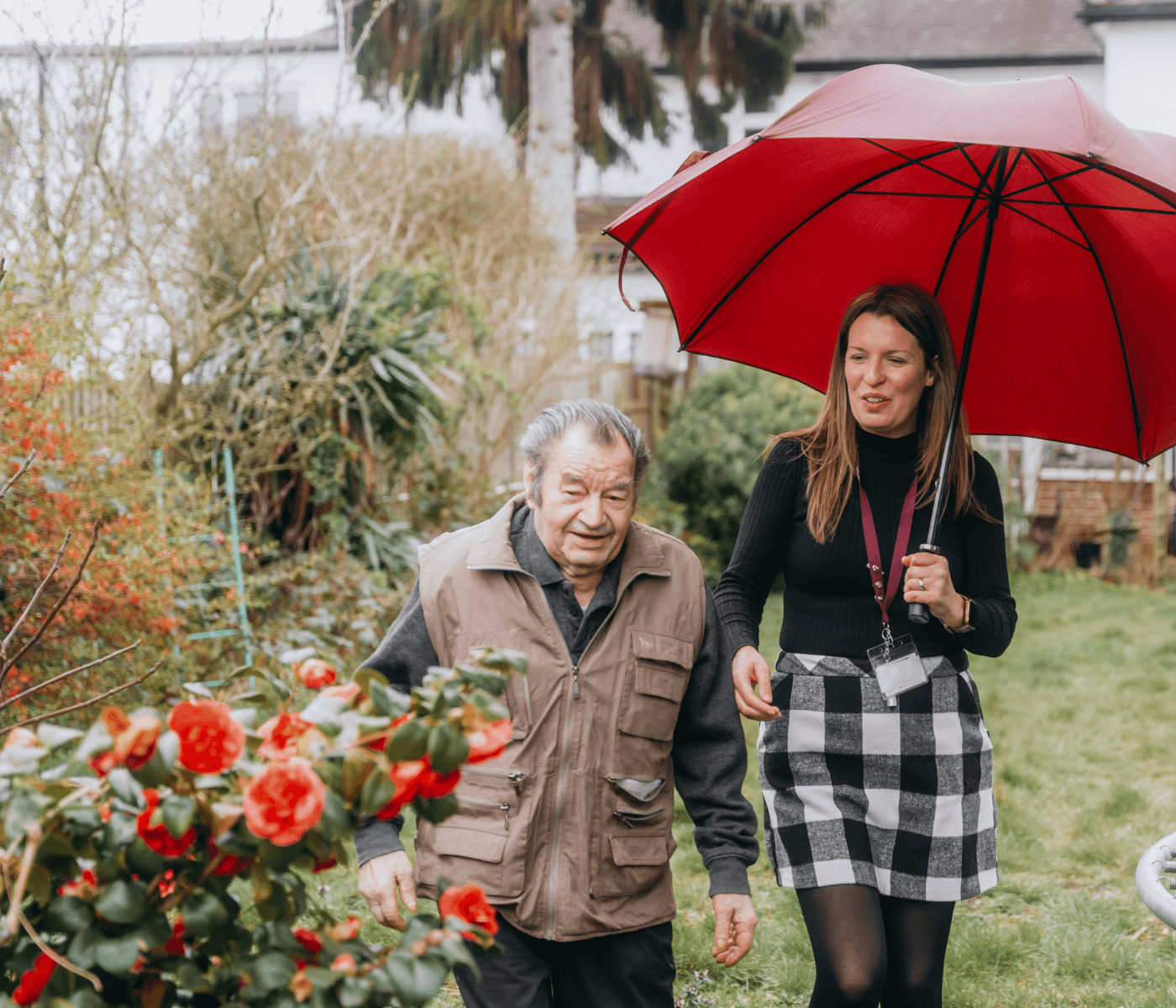 A woman holding a red umbrella walks with an elderly man in a garden filled with vibrant flowers. - Home Instead