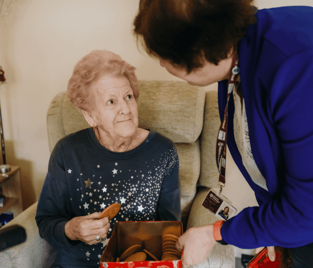 Elderly woman with a tin of cookies looks up at another woman standing beside her. - Home Instead