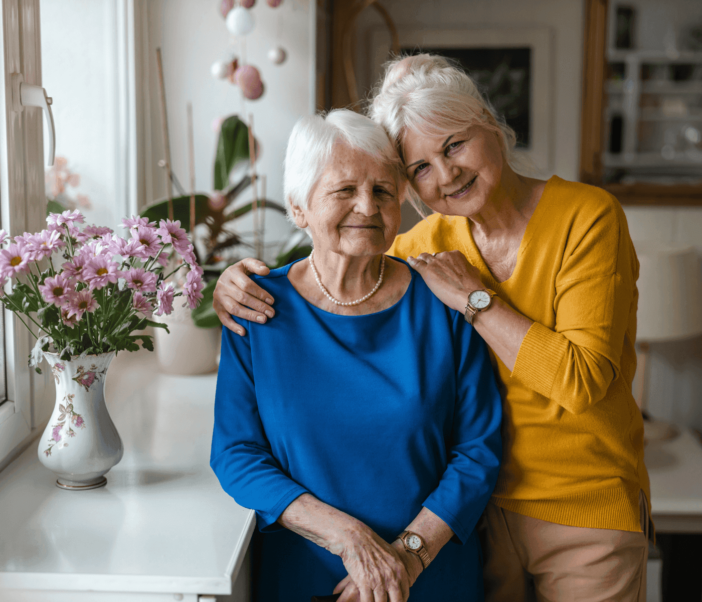 Two elderly women smiling, one in a blue dress and the other in a yellow sweater, standing next to pink flowers indoors. - Home Instead