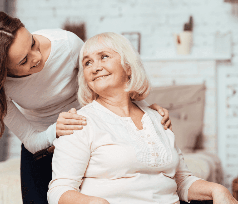 A young woman leans over, smiling at an elderly woman seated in a wheelchair, both looking at each other happily. - Home Instead