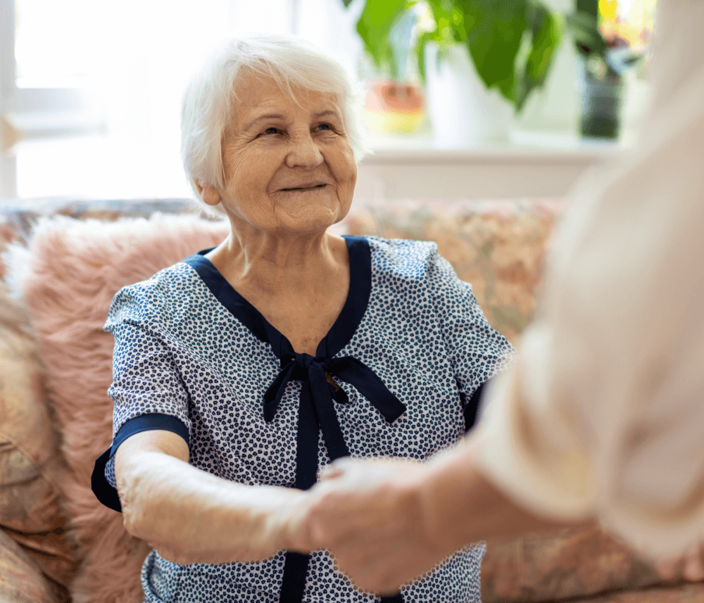 Elderly woman smiling and holding hands with a caregiver in a cozy living room setting with plants in the background. - Home Instead