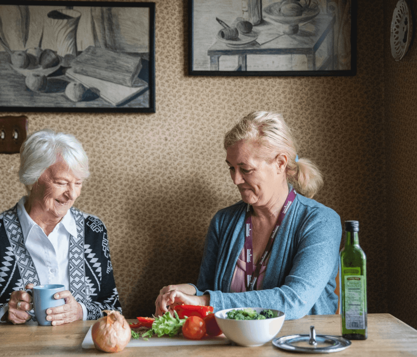 Two women sit at a kitchen table with vegetables, smiling, one holding a mug and the other preparing food. - Home Instead