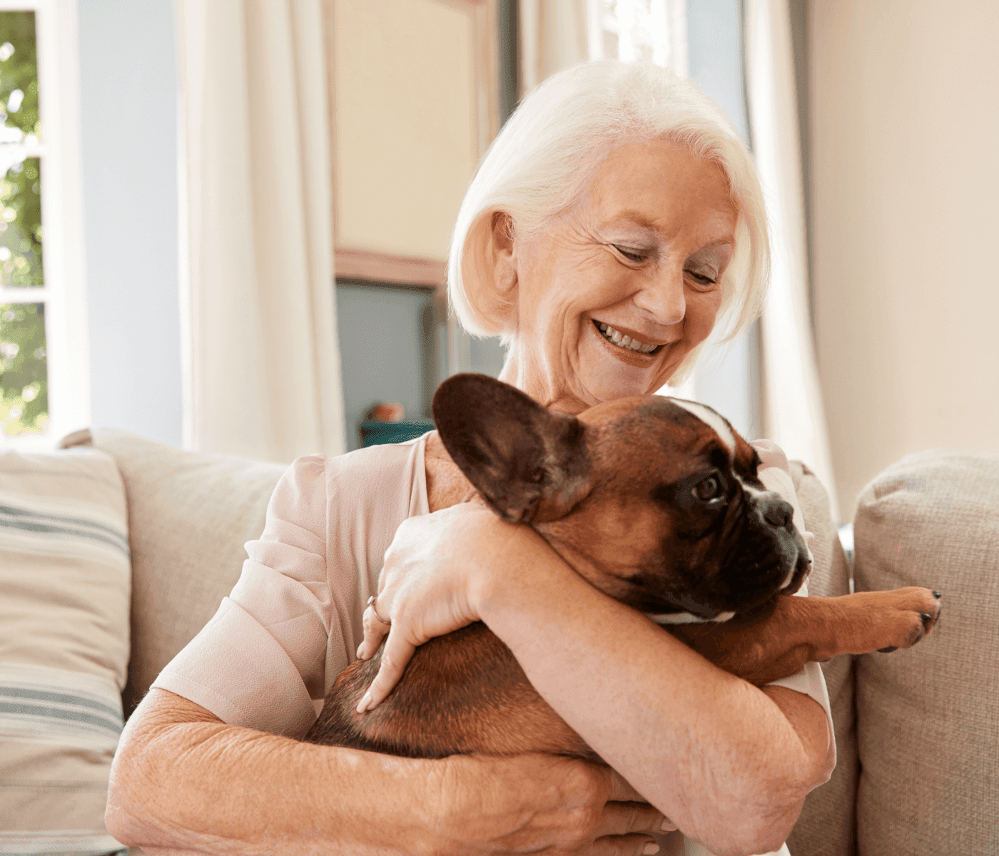 Smiling elderly woman hugging a brown French Bulldog on a couch in a bright living room. - Home Instead