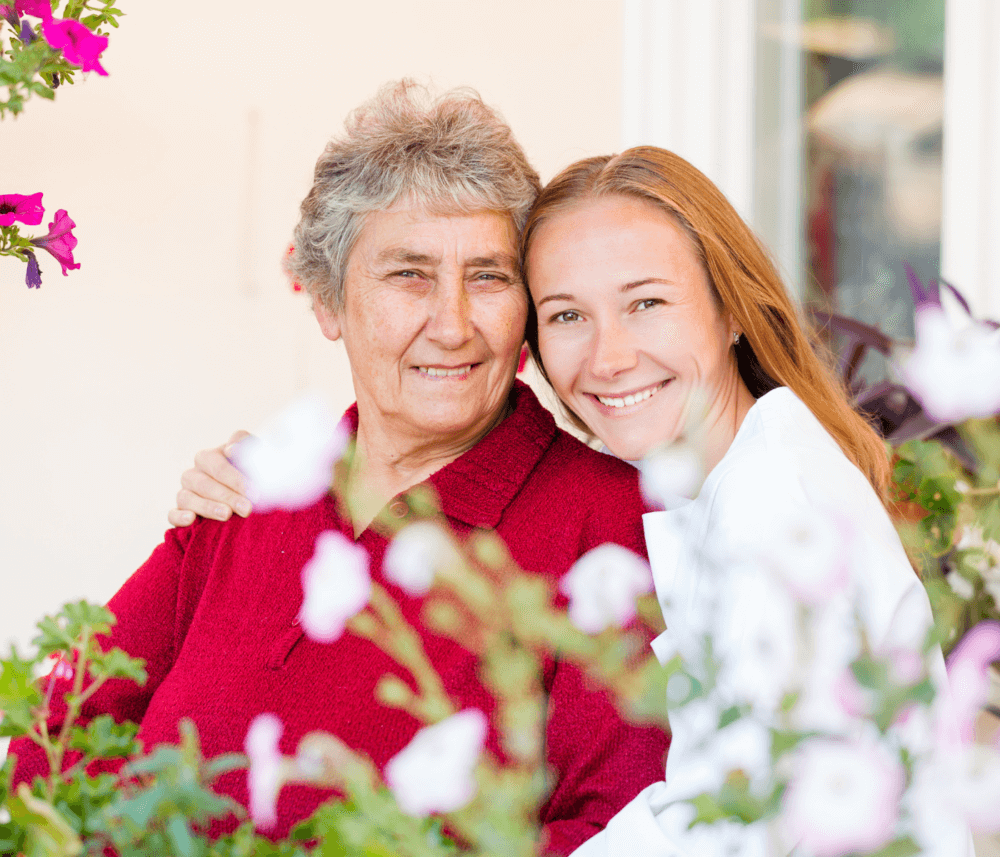 An elderly woman in a red sweater and a younger woman in white embrace, smiling amidst blooming flowers. - Home Instead
