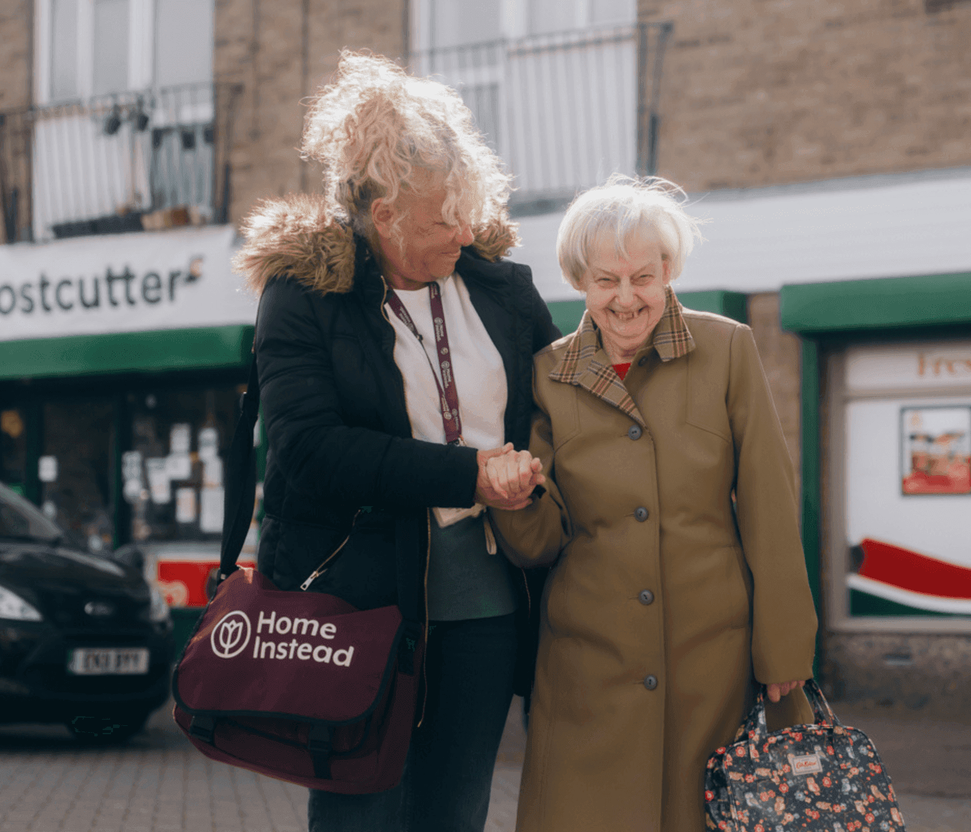A caregiver with a Home Instead bag helps a smiling elderly woman walk outside in a town setting. - Home Instead
