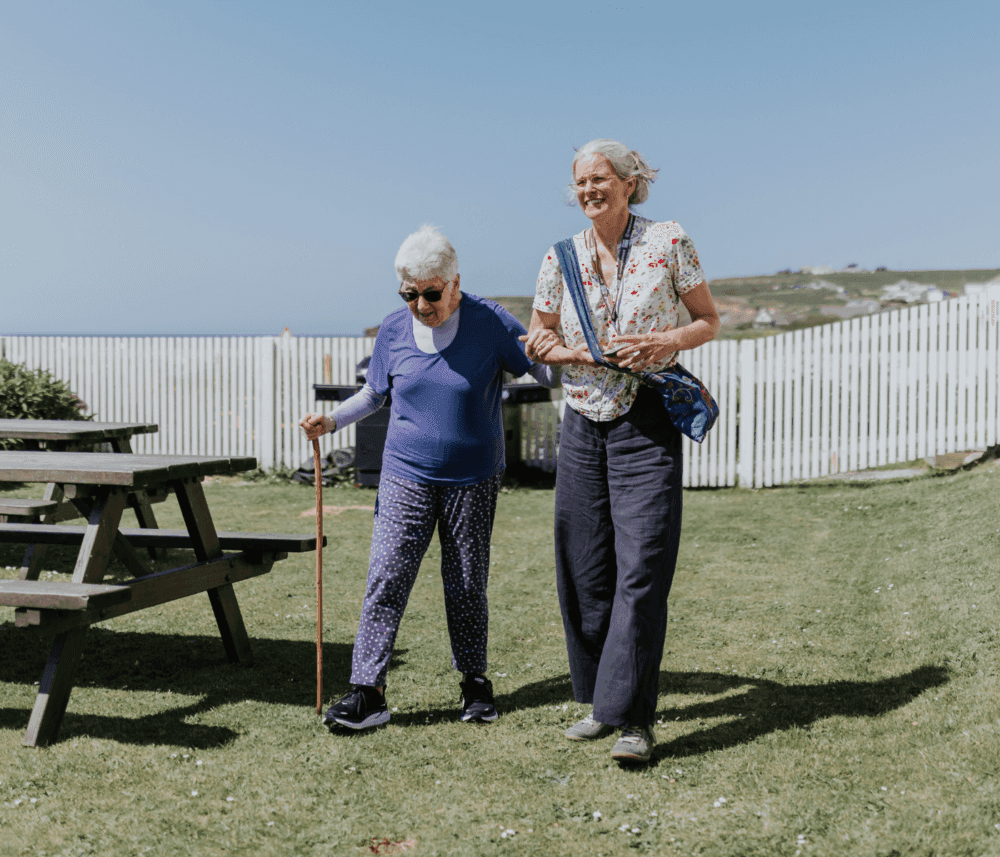 Two elderly women walking outdoors, one using a cane and the other providing support. A fence and picnic table are nearby. - Home Instead