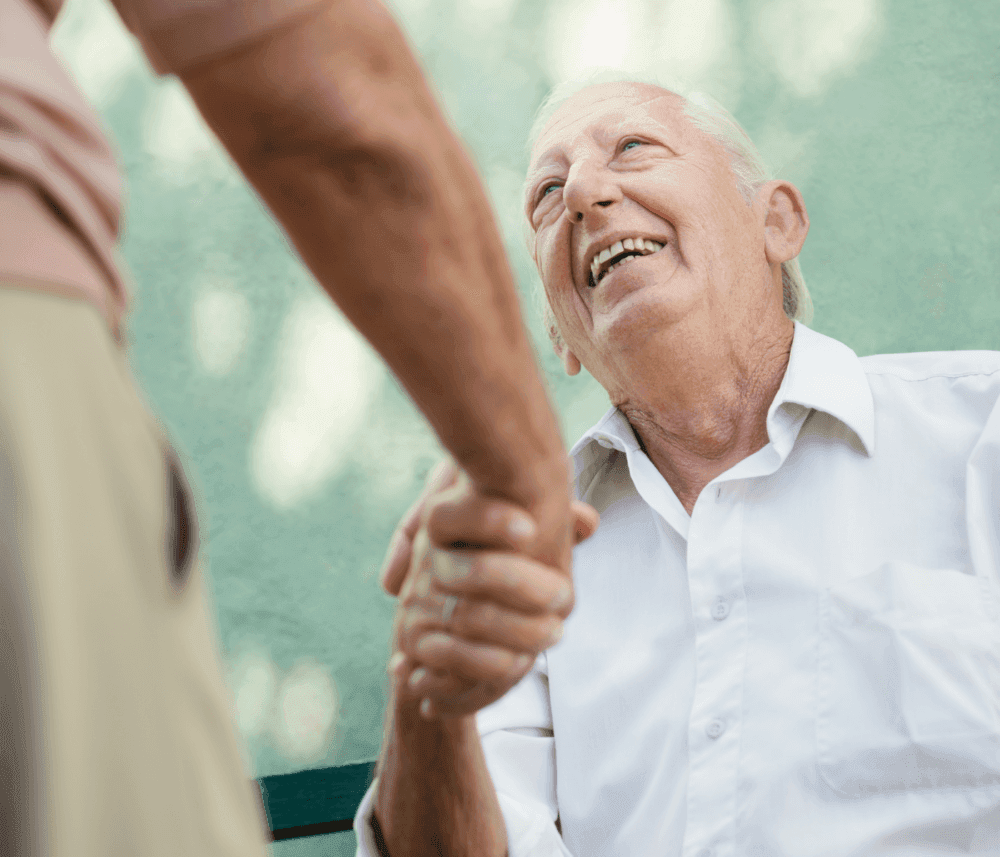 Elderly man in a white shirt smiling warmly while shaking hands with another person outdoors. - Home Instead