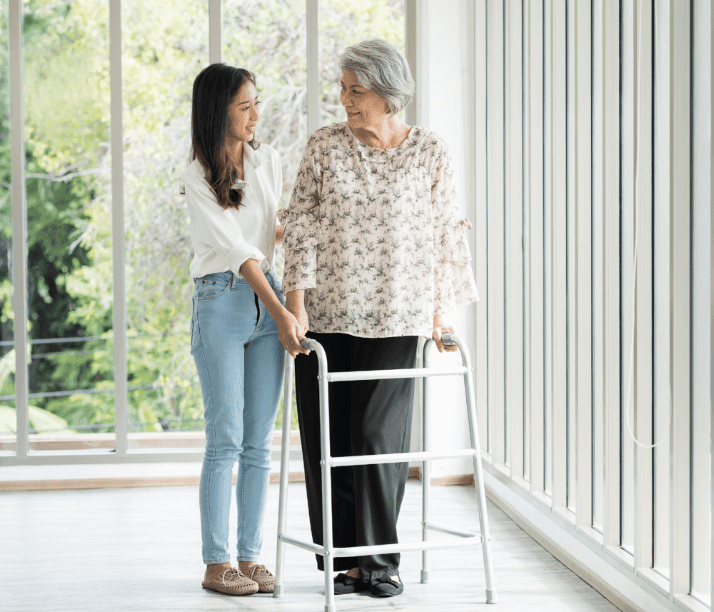 A young woman assists an elderly woman using a walker in a bright room with large windows and greenery outside. - Home Instead