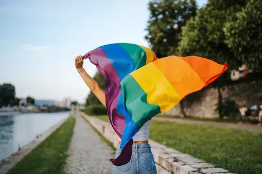Person waving a rainbow flag near a riverside path with trees and a stone wall in the background. - Home Instead