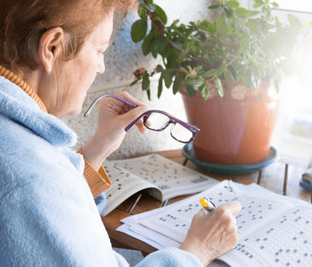 A person with glasses doing a crossword puzzle by a window with a potted plant nearby. - Home Instead