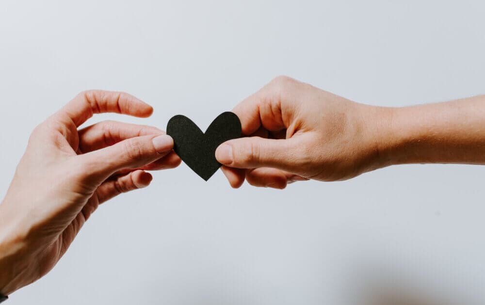 Two hands holding a black paper heart together against a plain white background. - Home Instead