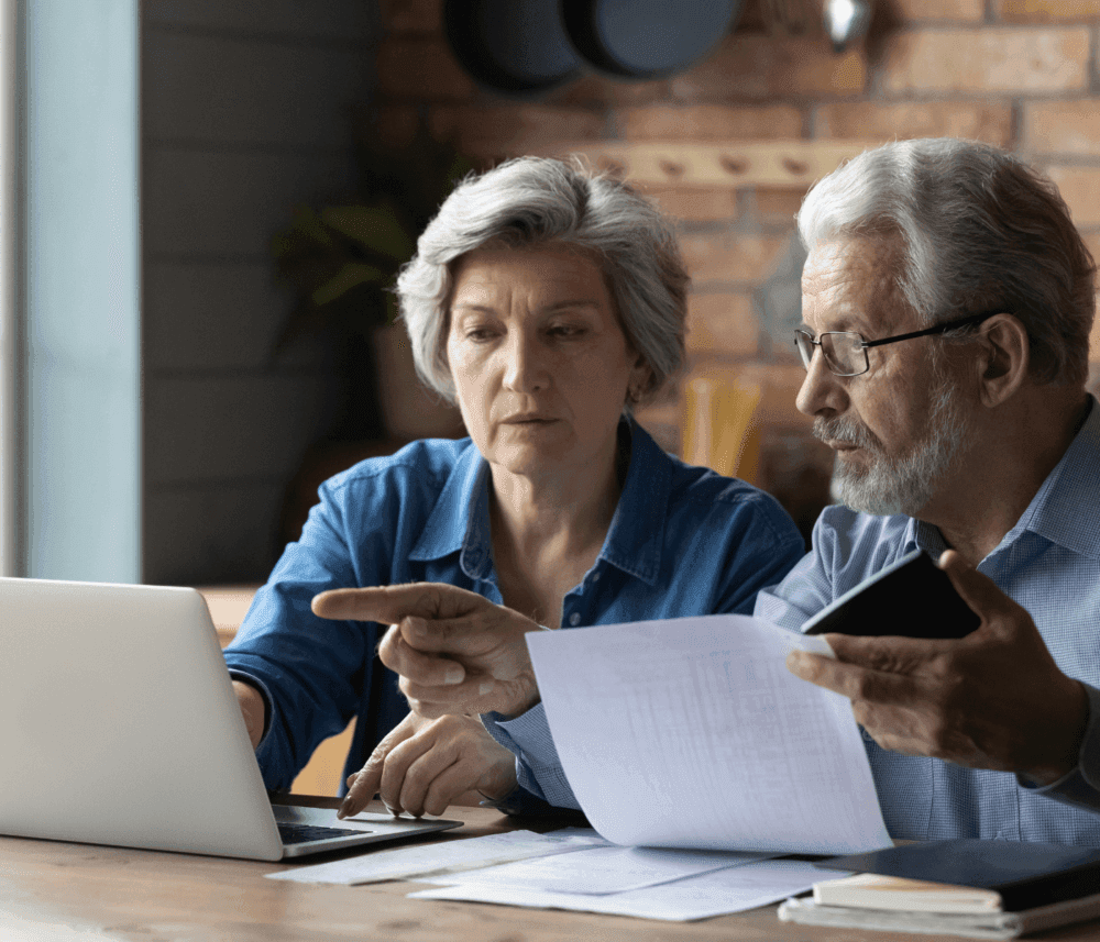 Elderly couple at a table, reviewing documents and working on a laptop together. - Home Instead