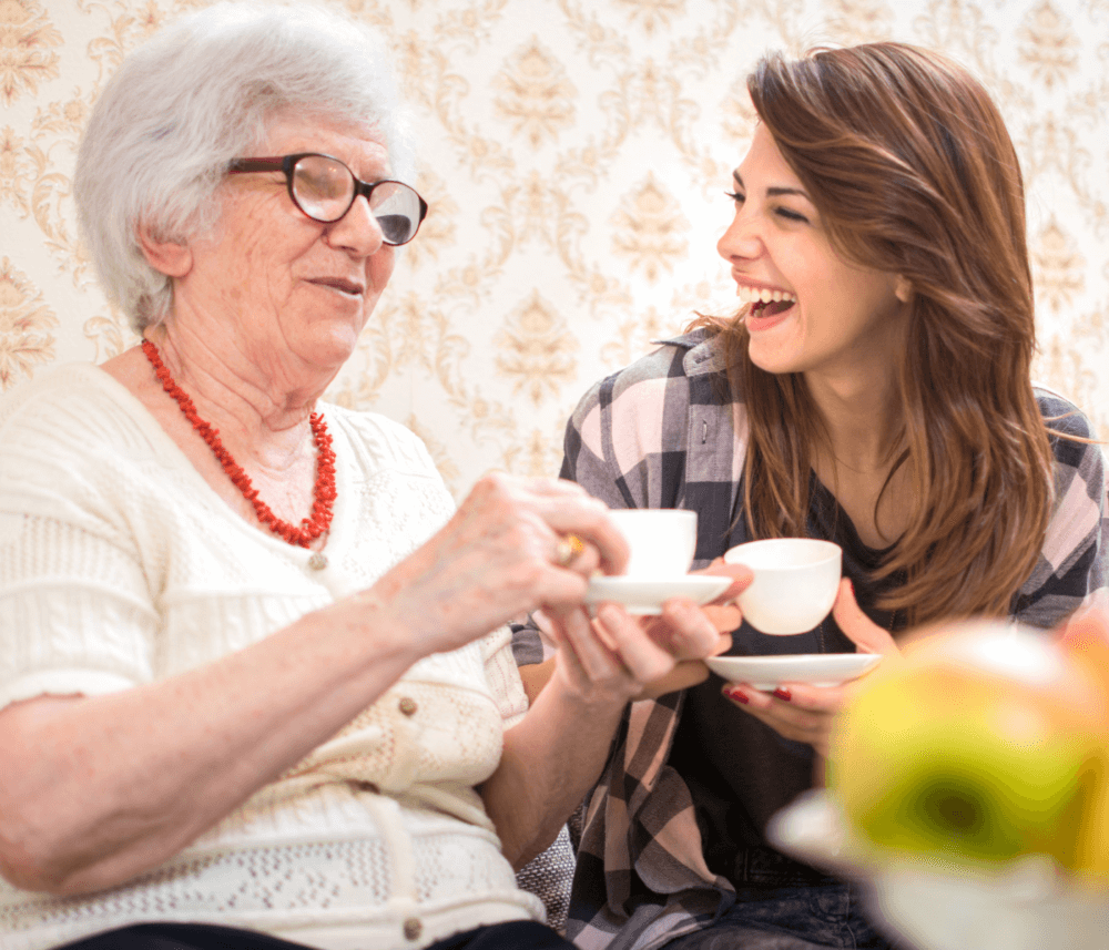 An elderly woman and a young woman enjoying tea together, both smiling and sitting on a couch. - Home Instead