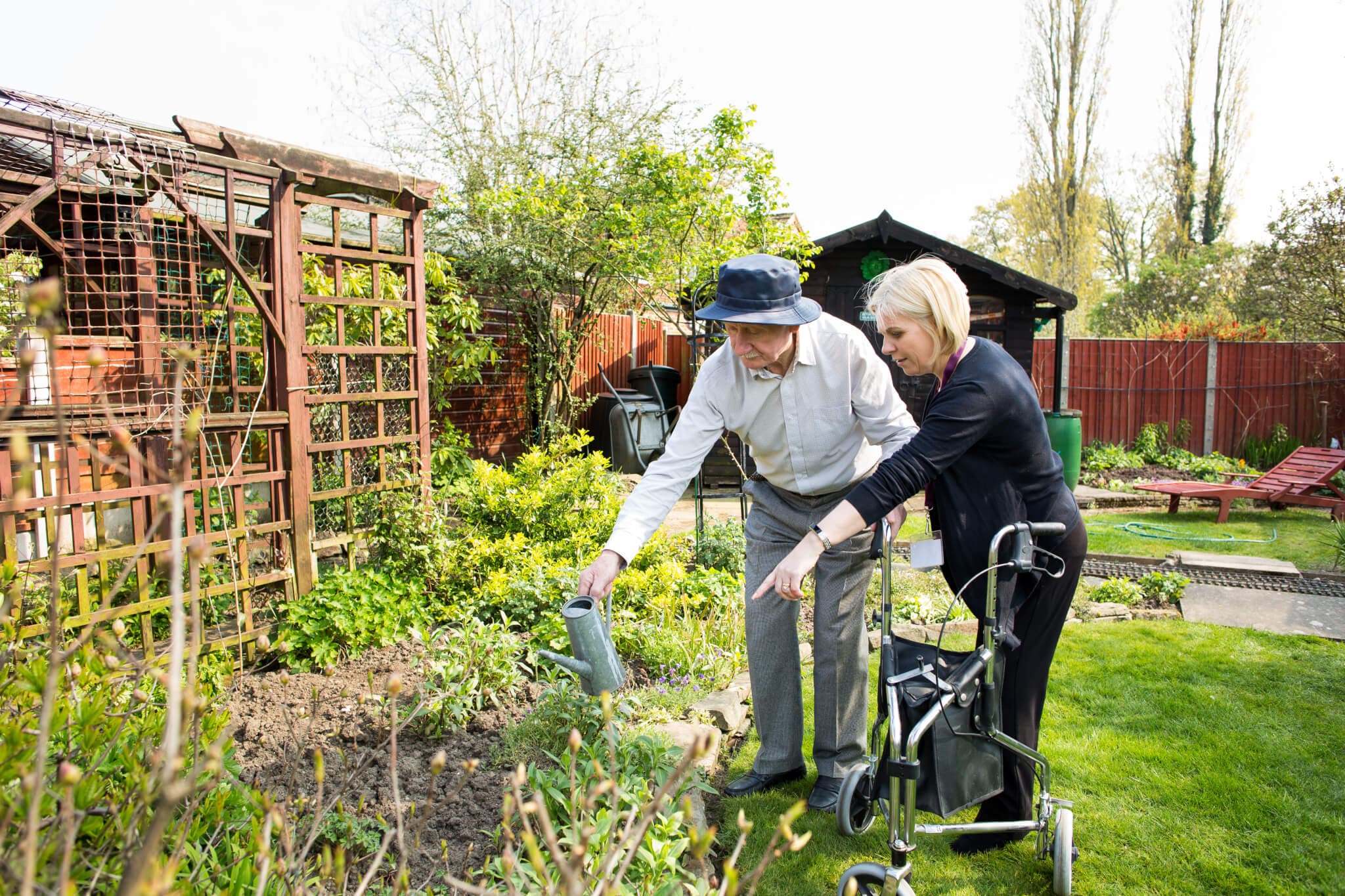 Two people gardening together in a backyard, one with a walker and the other watering the plants. - Home Instead