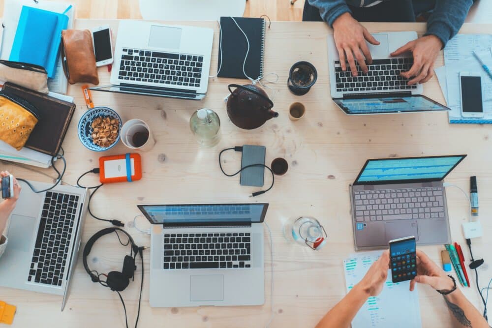 Top view of a cluttered desk with laptops, notebooks, headphones, snacks, and people using various electronic devices. - Home Instead