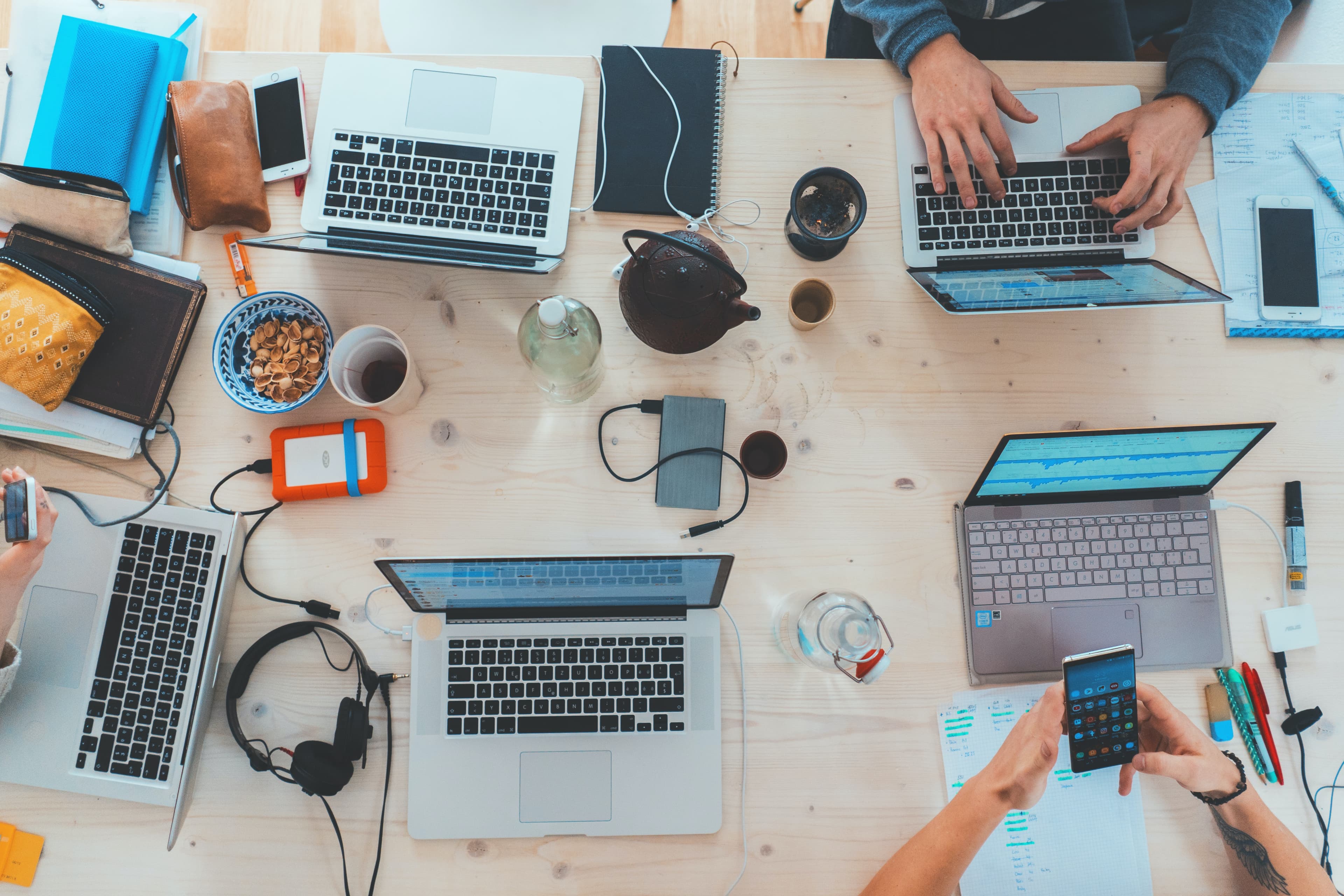 Top view of a cluttered desk with laptops, notebooks, headphones, snacks, and people using various electronic devices. - Home Instead