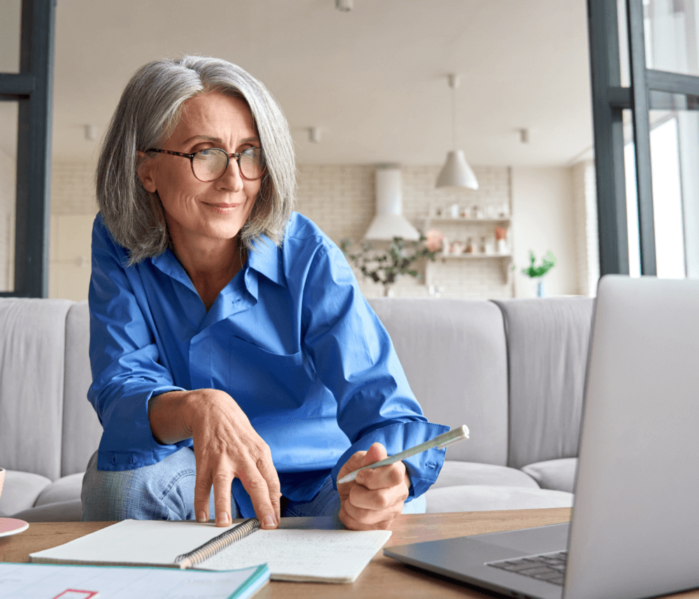 Older woman with gray hair and glasses, writing in a notebook, seated on a sofa, with an open laptop in front of her. - Home Instead