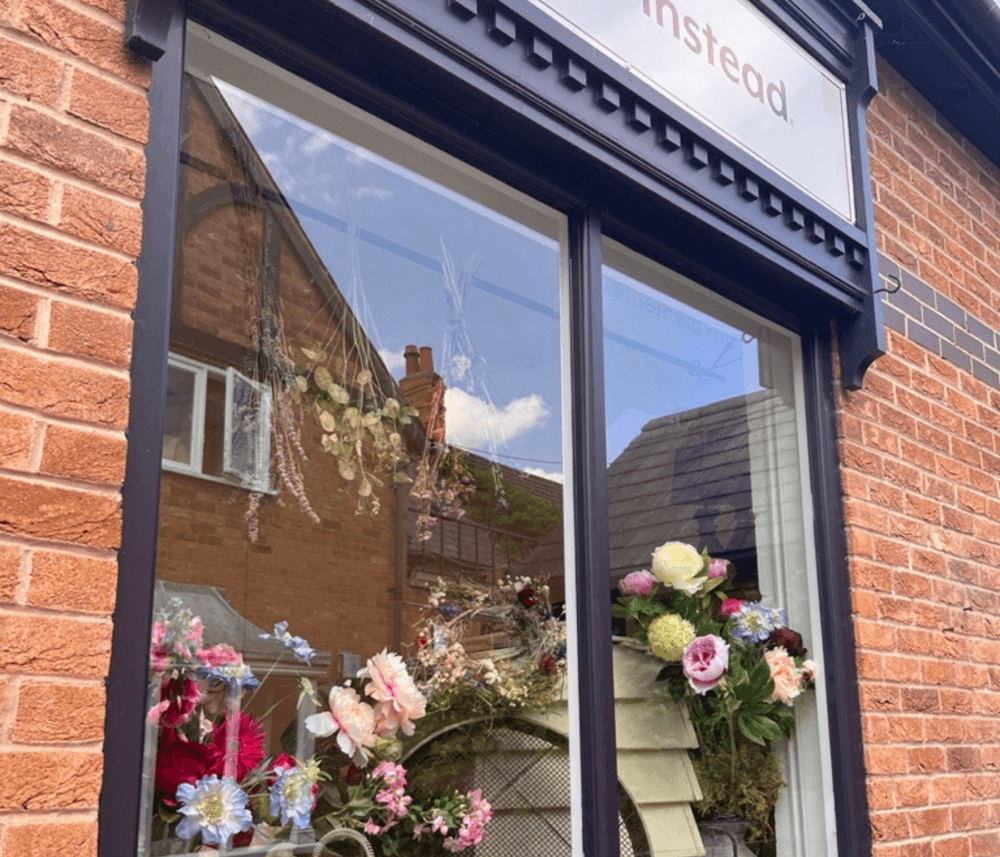 A flower shop window display with a mix of dried and fresh flowers against a brick wall, reflecting nearby buildings. - Home Instead