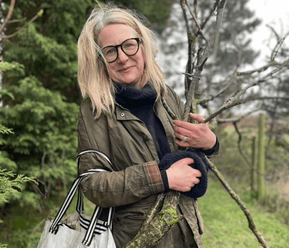 A person with glasses and a coat hugs a tree branch outdoors, holding a white bag with black stripes. - Home Instead