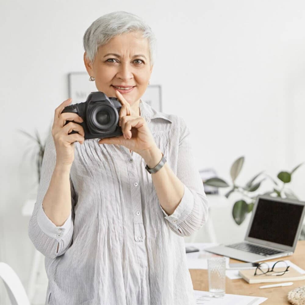 A woman with short gray hair, holding a camera, stands in a bright room with a laptop and plants in the background. - Home Instead
