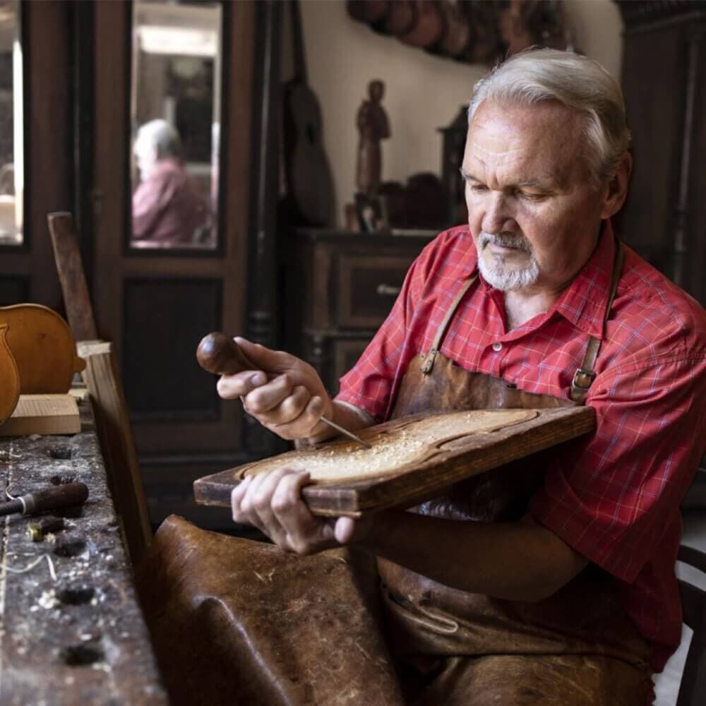 An older man in a workshop carves wood with a chisel, wearing a red shirt and brown apron. Tools and wood shavings are on the table. - Home Instead