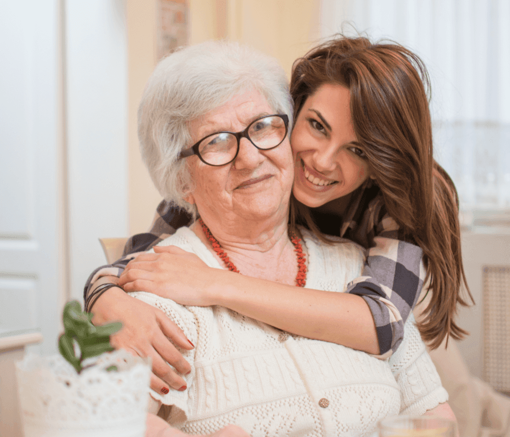 A young woman hugs an elderly woman who is wearing glasses and a white sweater, both smiling warmly at the camera. - Home Instead