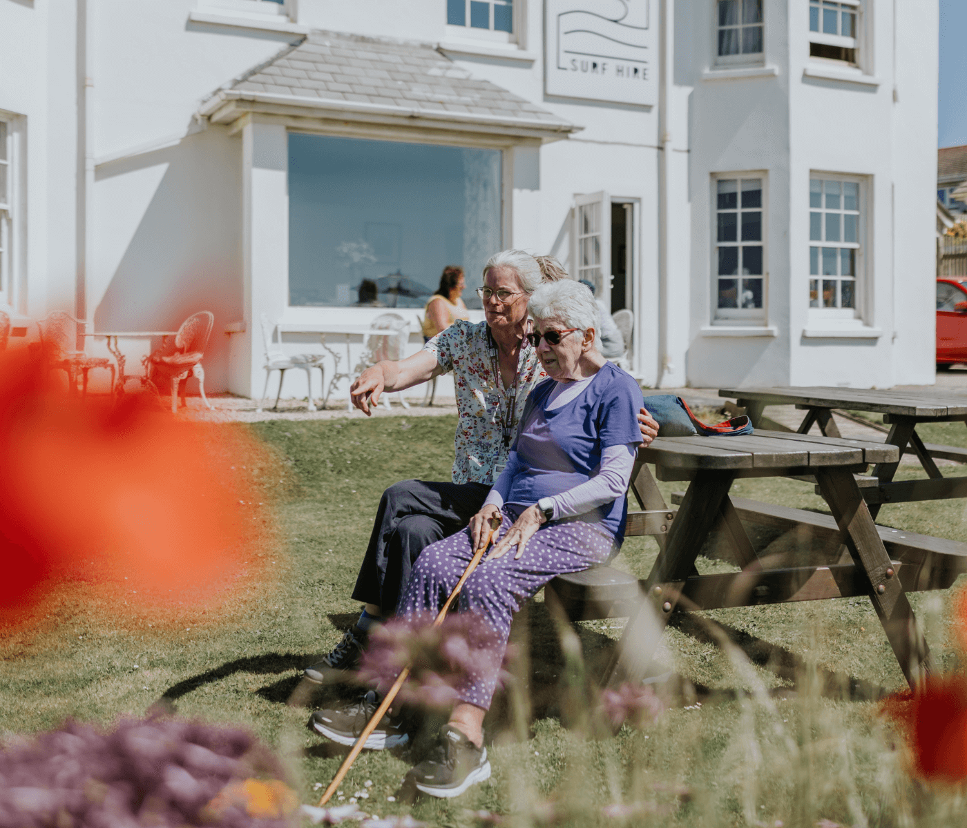 Two elderly women sitting on a bench outside a white building with flowers in the foreground. - Home Instead