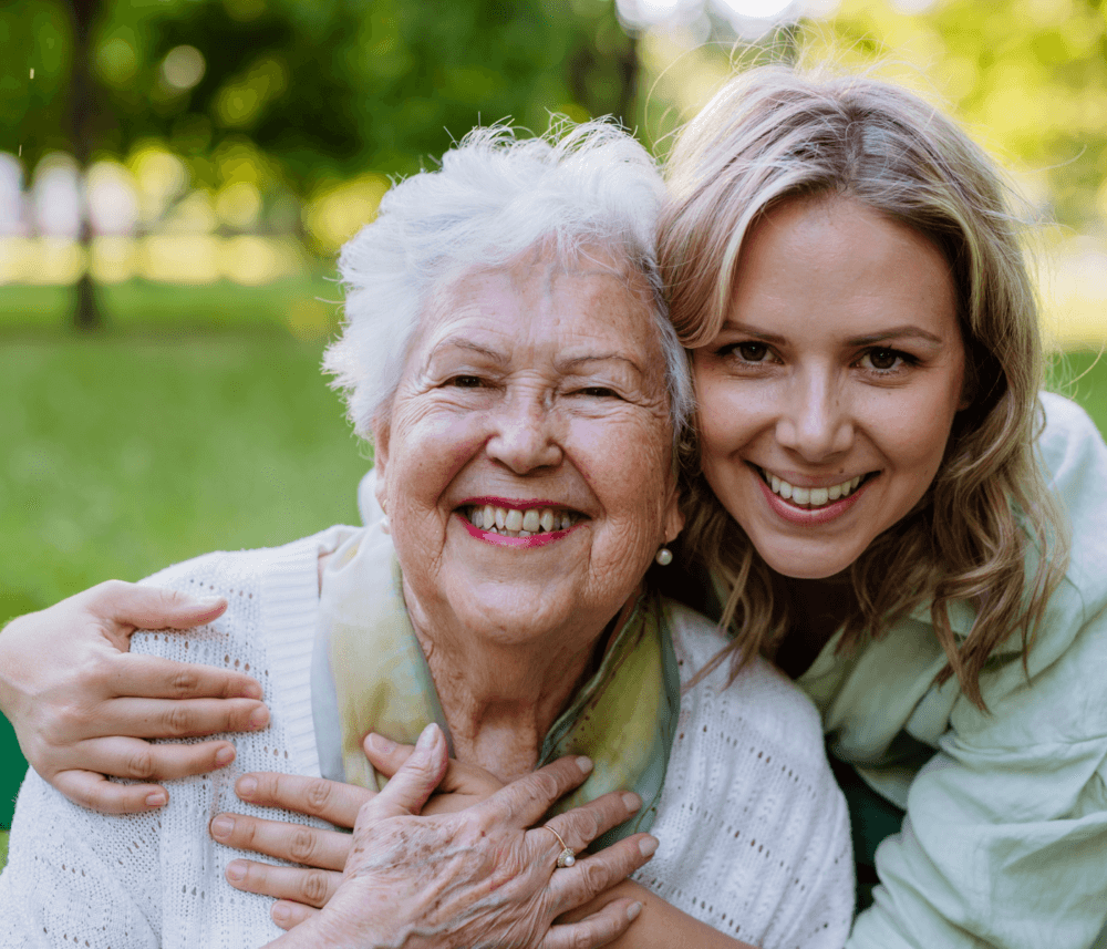 A smiling older woman and a younger woman embracing outdoors with greenery in the background. - Home Instead