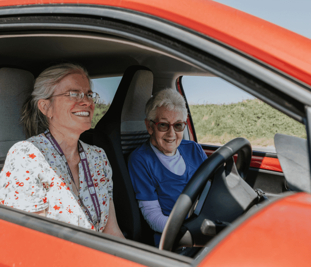 Two elderly women smiling while sitting inside a red car, with one in the driver's seat. - Home Instead