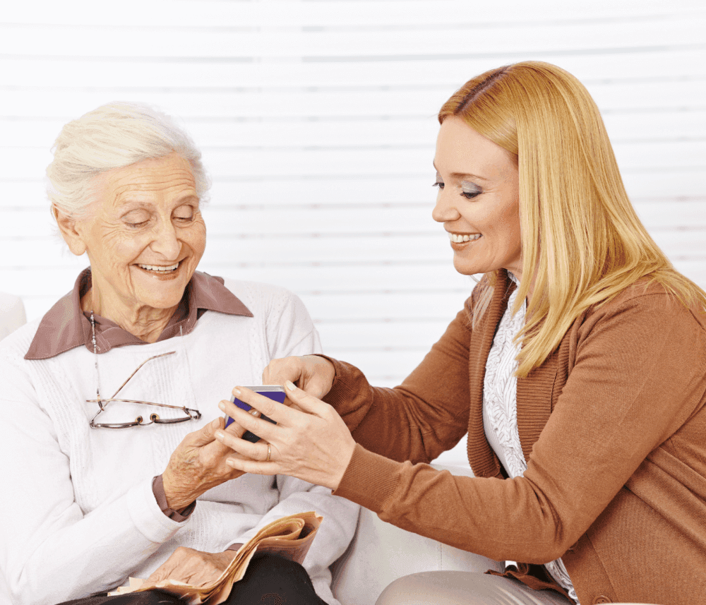 An elderly woman smiles while a younger woman hands her a cup. Both are seated, and a pair of glasses rests on the elder's sweater. - Home Instead