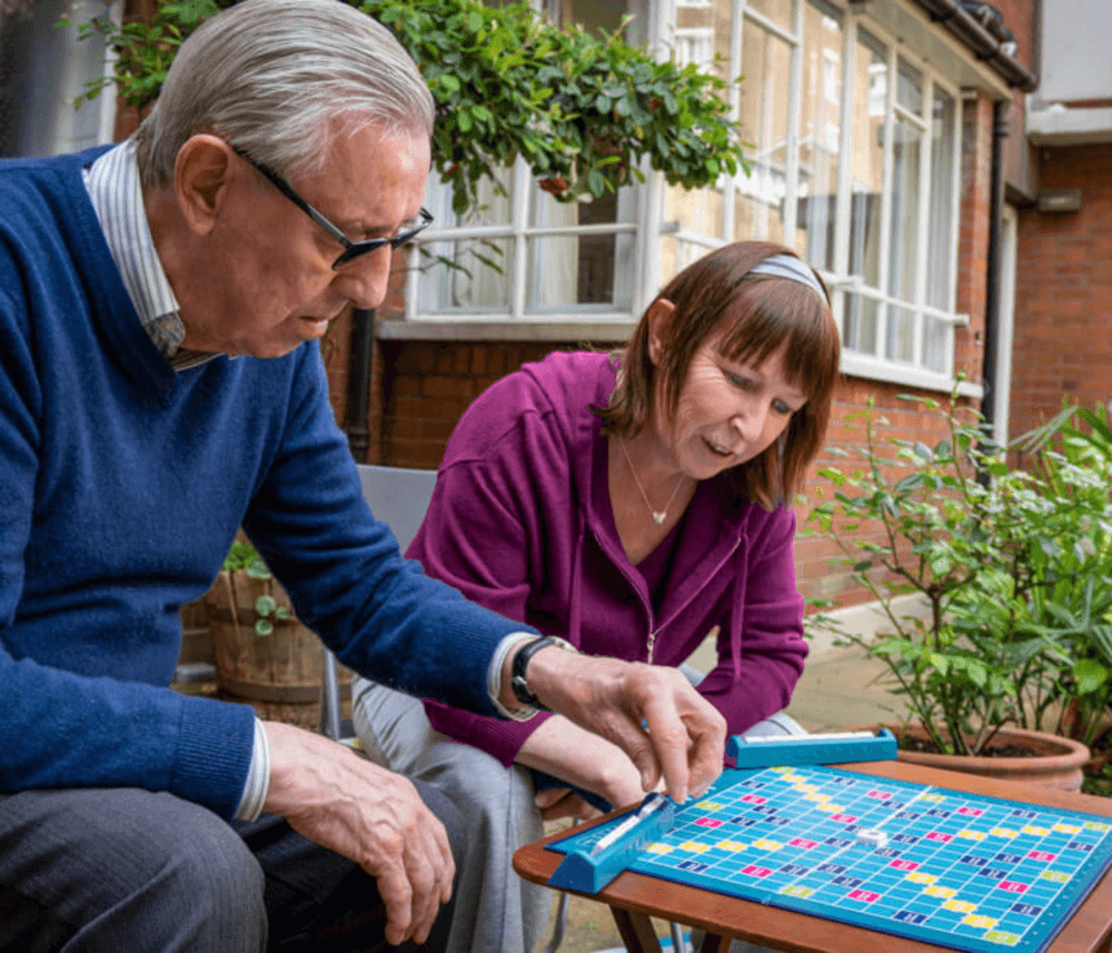 Two people playing Scrabble on a table outside, surrounded by greenery and buildings in the background. - Home Instead