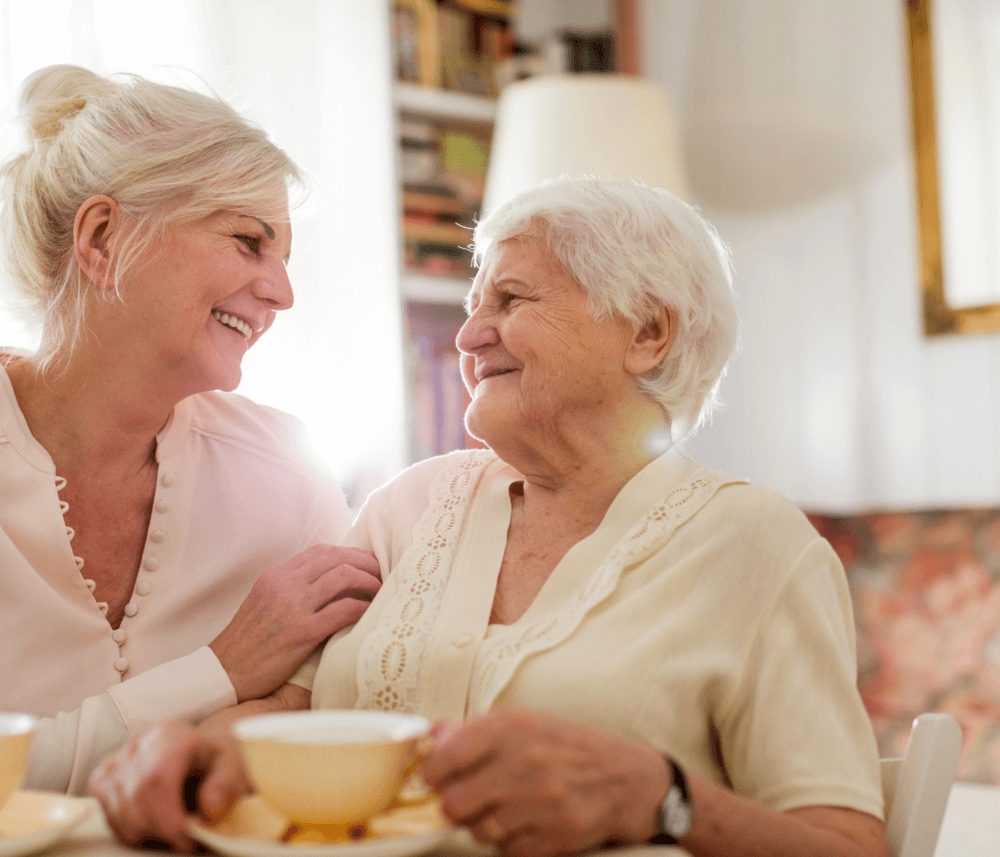 Two elderly women sitting together, smiling and holding teacups in a cozy, well-lit room. - Home Instead