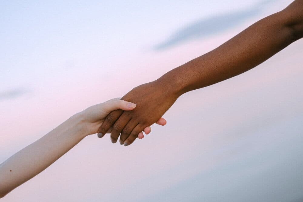 Two hands, one light-skinned and one dark-skinned, clasp each other against a serene background of a pastel sky. - Home Instead