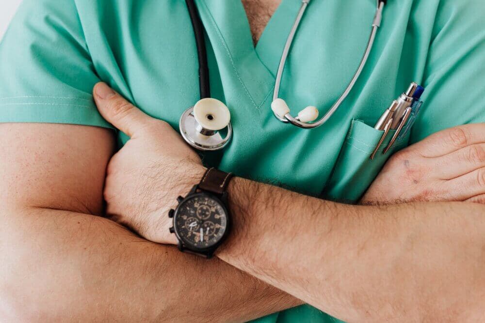 A person in green scrubs with folded arms, wearing a stethoscope, a wristwatch, and pens in the pocket. - Home Instead